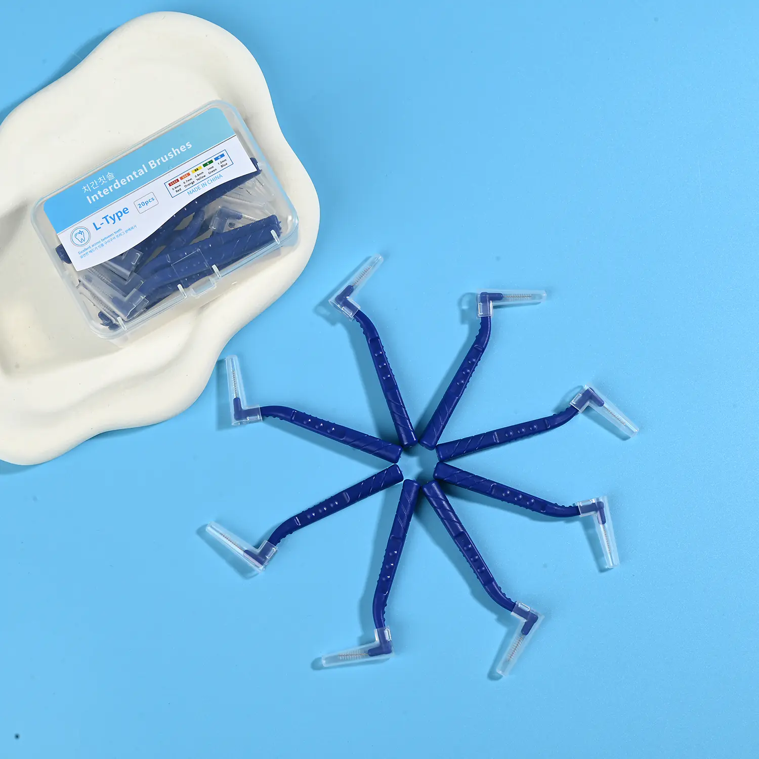 Dark blue L-type interdental brushes arranged in radial star shape with clear plastic storage box on beige irregular pad and light blue background