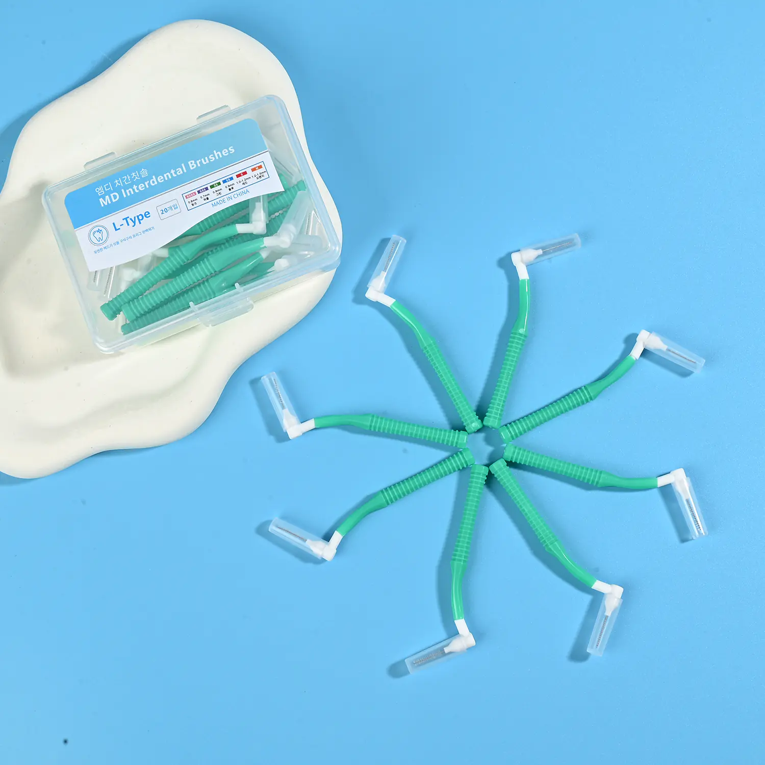 Green L-type MD interdental cleaners arranged in radial star shape alongside clear plastic storage box on beige pad and light blue background
