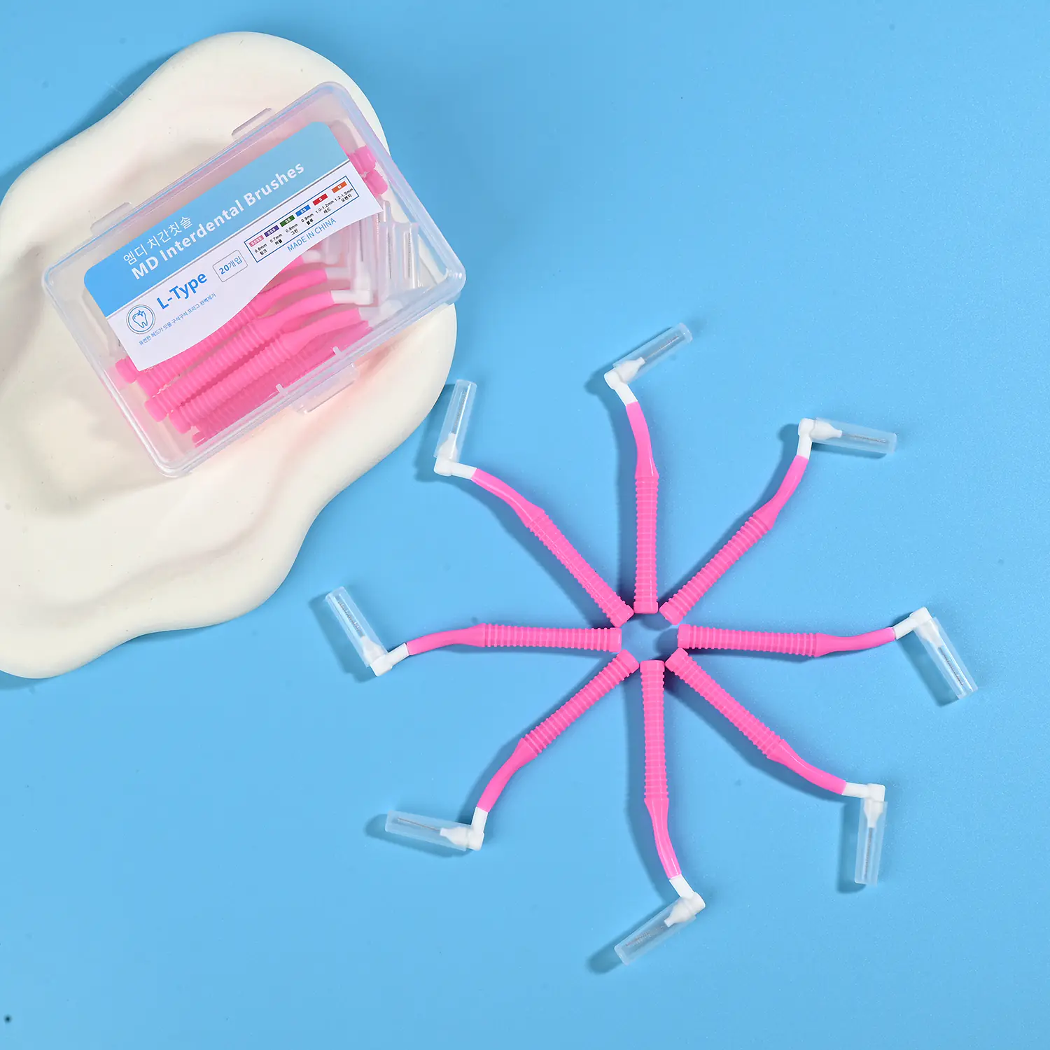 Pink L-type MD interdental cleaners arranged radial star shape with original clear plastic box on beige irregular pad and light blue background