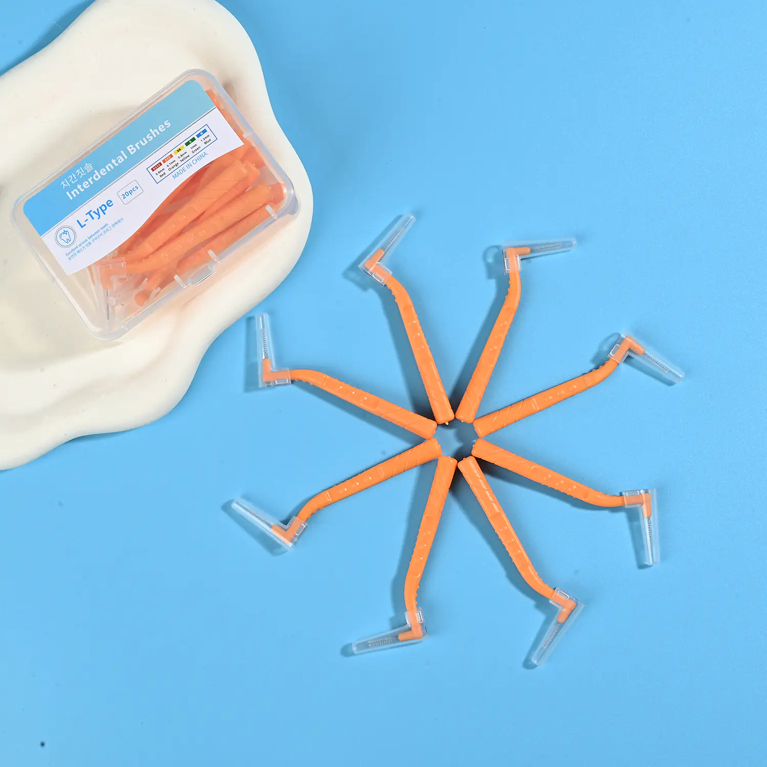Orange L-type interdental cleaners arranged radial star shape alongside original clear plastic box on irregular beige pad and bright blue background