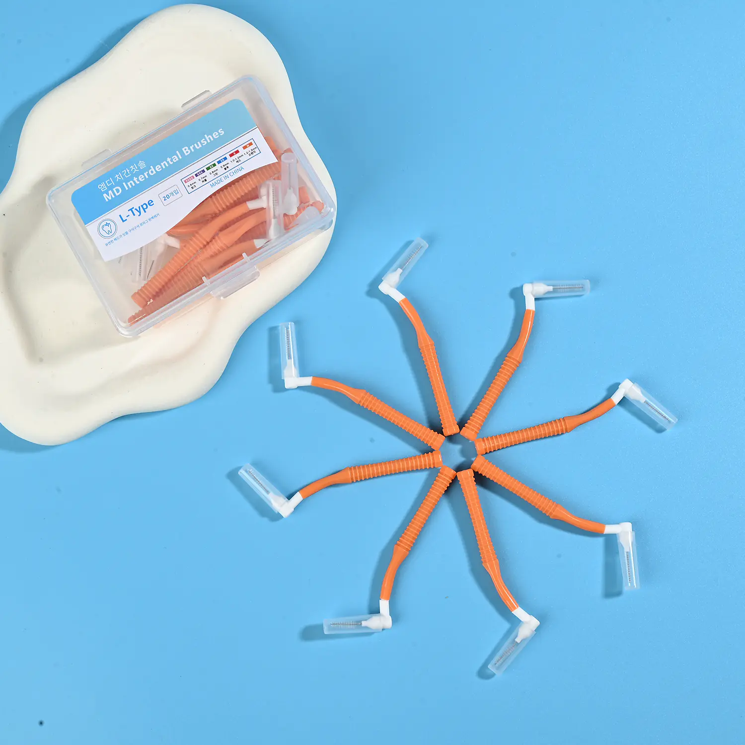 Orange L-type MD interdental brushes arranged in star shape with clear plastic storage box on beige pad and blue background